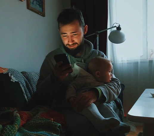 Father sitting in a dimly lit room while holding his baby and reading messages on a smartphone, representing secure co parenting communication with Parent Link.