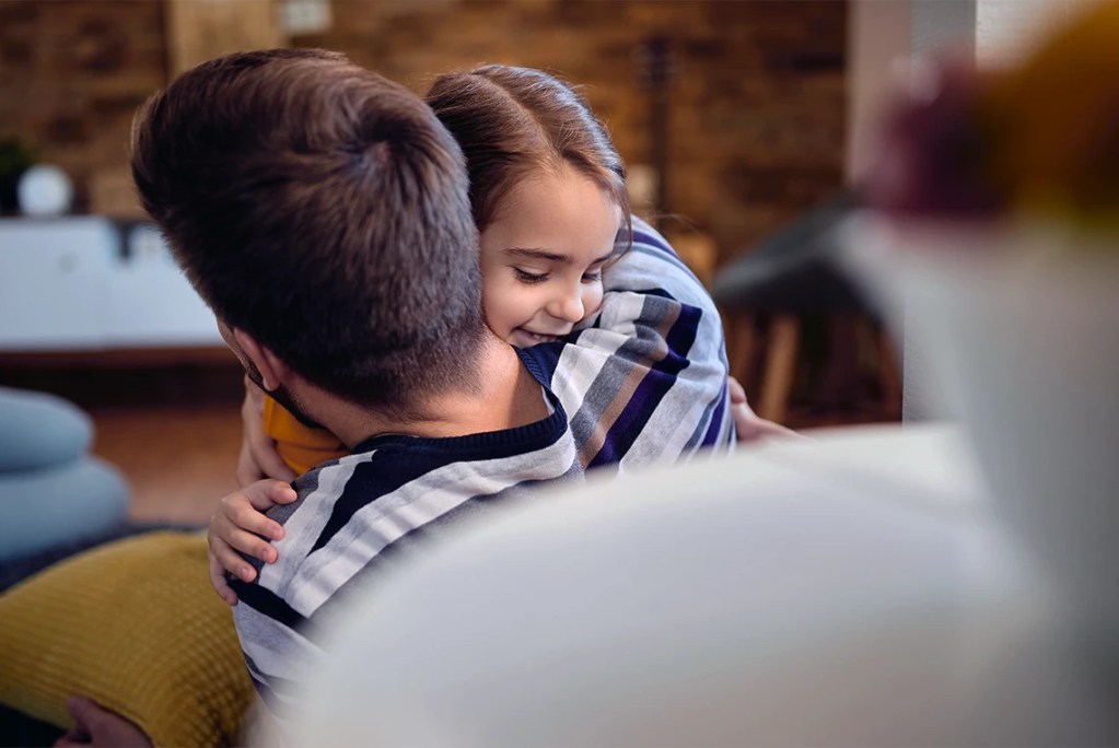 A young girl smiling and hugging her father in a warm home environment, illustrating the emotional stability and family history that Ari's Law protects through co-parenting data rights.