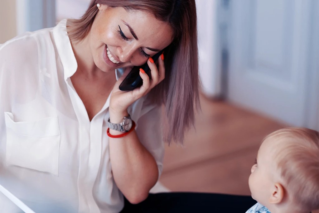 A smiling mother talking on a mobile phone while looking at her baby, illustrating the concept of managed communication in a professional co-parenting environment.