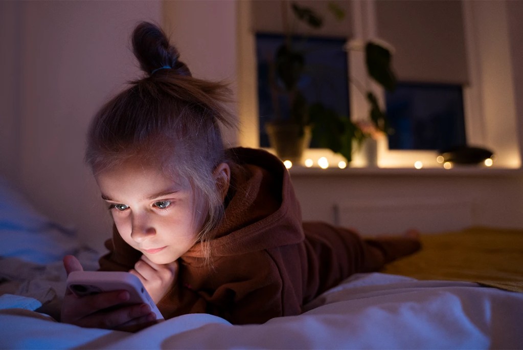 A young child lying in bed using a smartphone at night, illustrating the need for digital child safety and parental transparency.