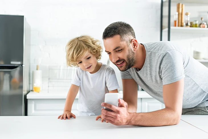 Father and young son looking at a smartphone in a bright kitchen while using the Parent Link co parenting app to manage schedules and messages.