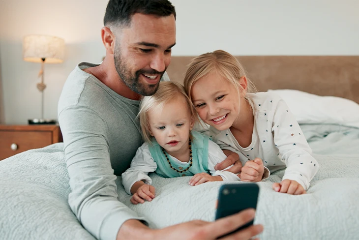 Father sitting on a bed with two daughters while using a smartphone that represents how Parent Link helps parents track custody time and coordinate family routines.