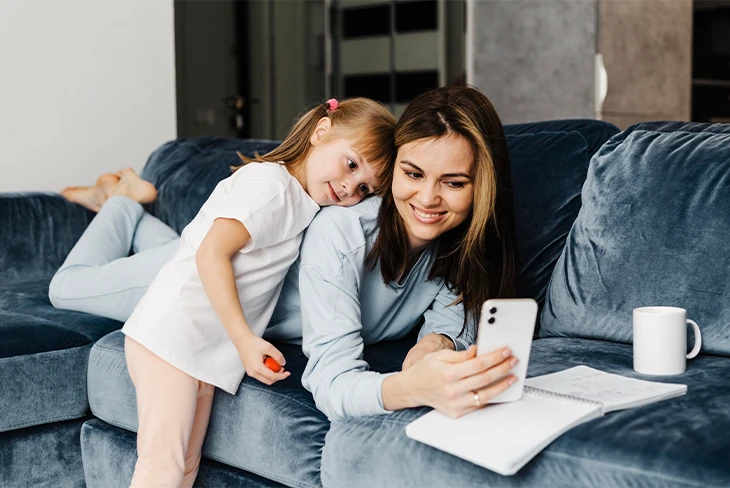 Mother and daughter smiling at a smartphone on a couch while using Parent Link to organize co parenting tasks and shared communication.