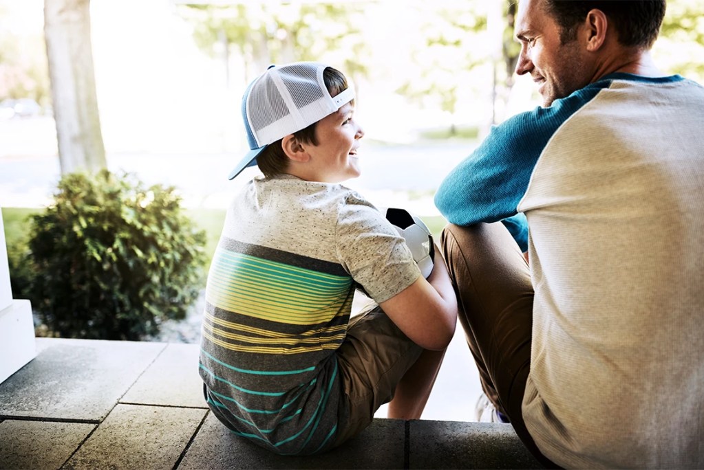 A father and son smiling while sitting on outdoor steps and talking, with a soccer ball between them, symbolizing positive co-parenting connection.