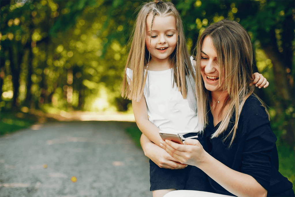 A mother and daughter looking at a phone together on a sunlit park path.