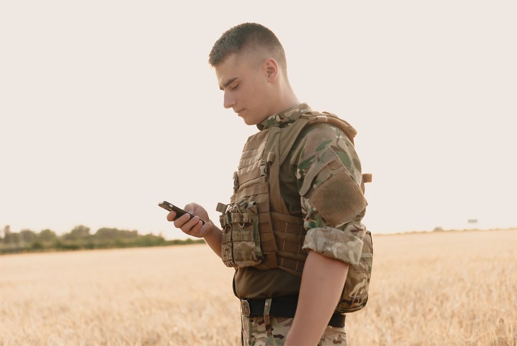 A military parent in uniform using a mobile device in a field to stay connected with their family during a deployment.